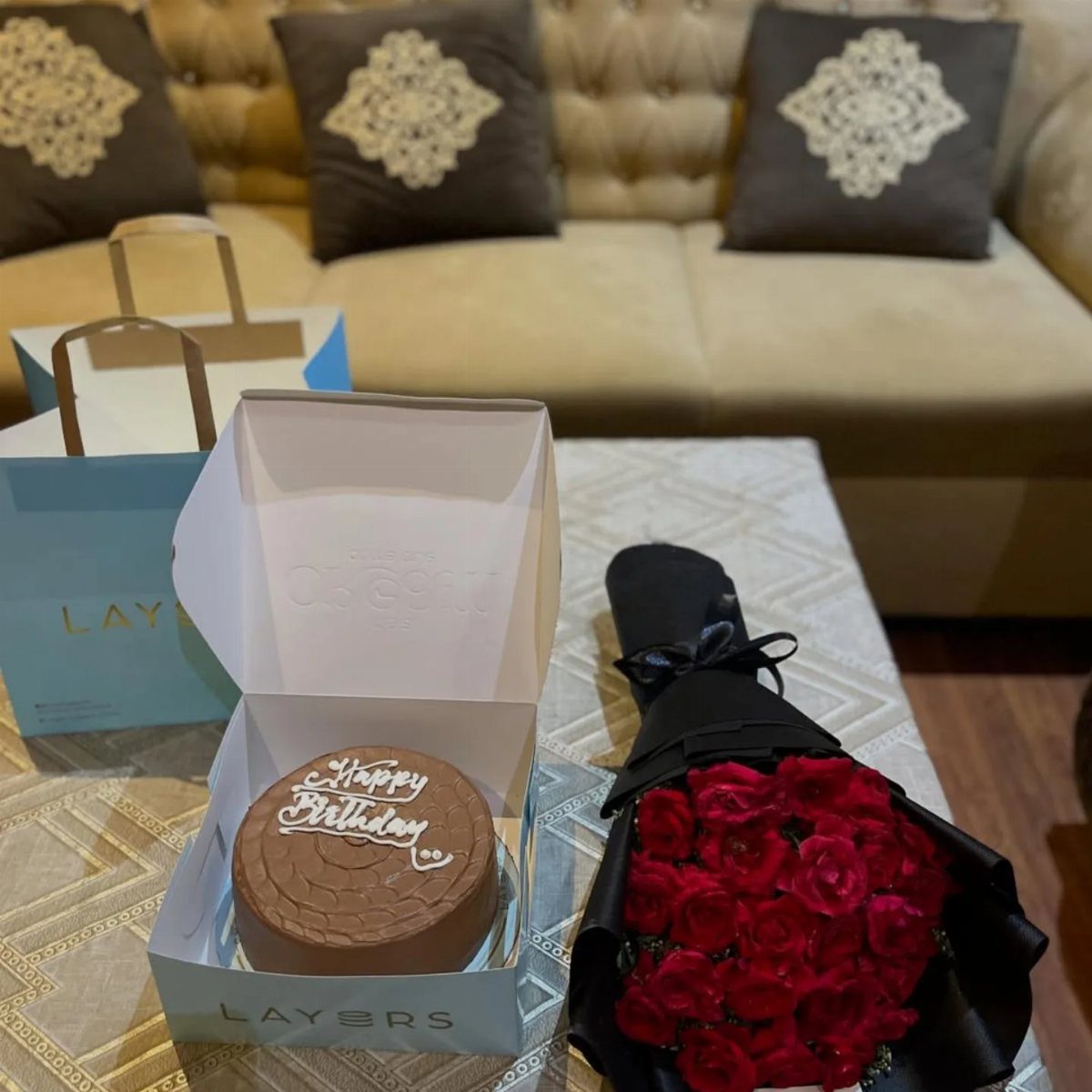 A chocolate birthday cake, red roses, and blue gift bags on a table.