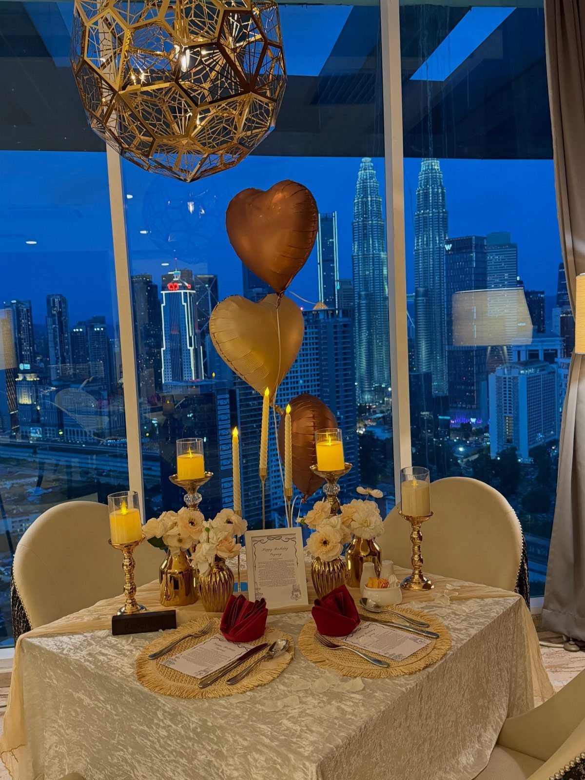 A romantic dinner table set for two with candles and heart balloons overlooking a city skyline.