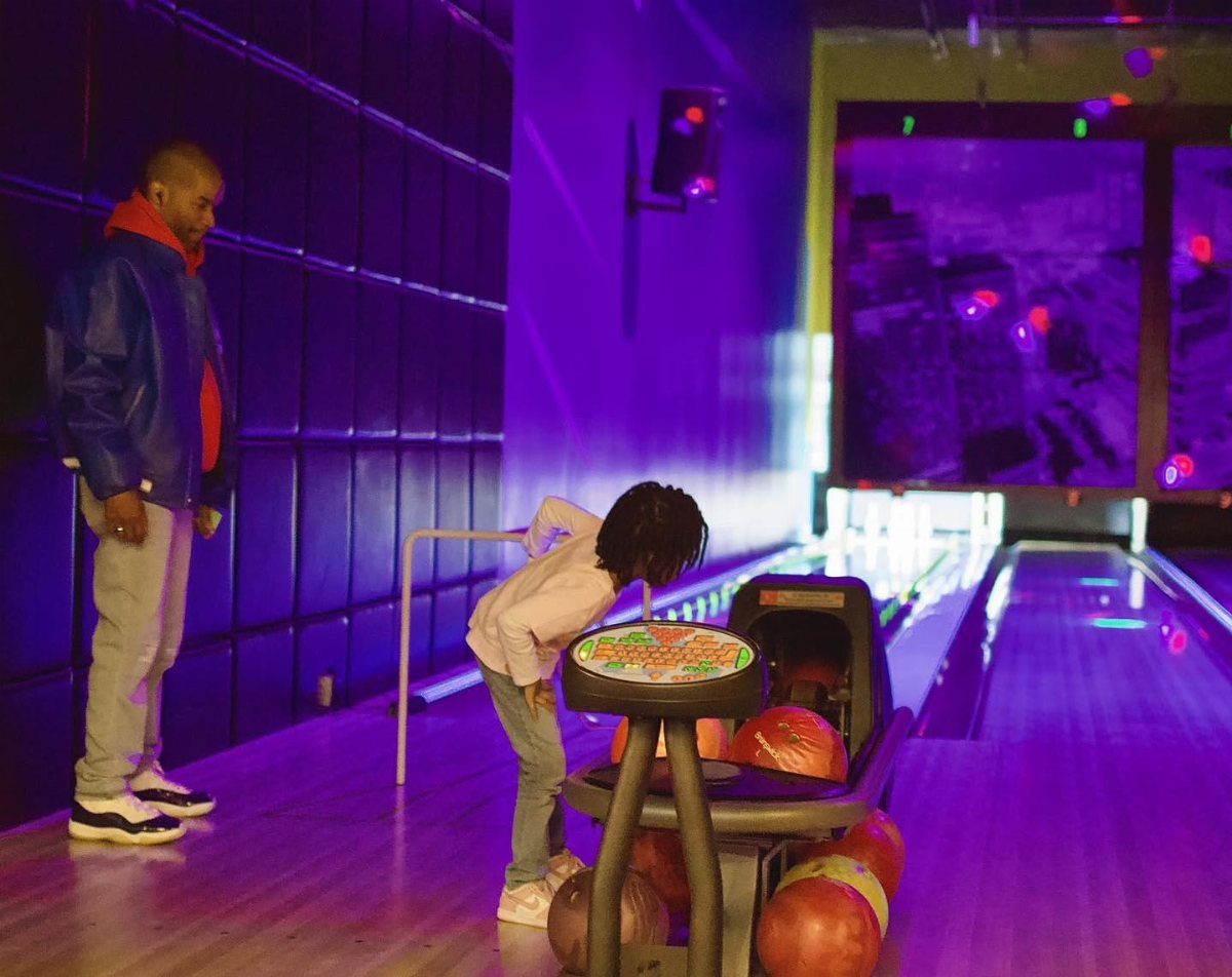 A man and a child leaning over a bowling ball return in a neon lit bowling alley.