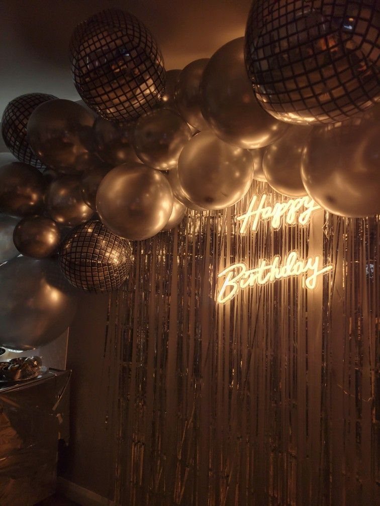 Silver disco balls and metallic balloons arranged above a shimmering fringe curtain with a neon Happy Birthday sign.