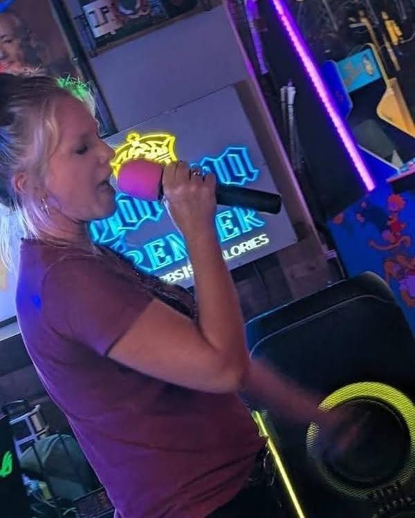 A young woman singing into a pink microphone under bright neon lights.