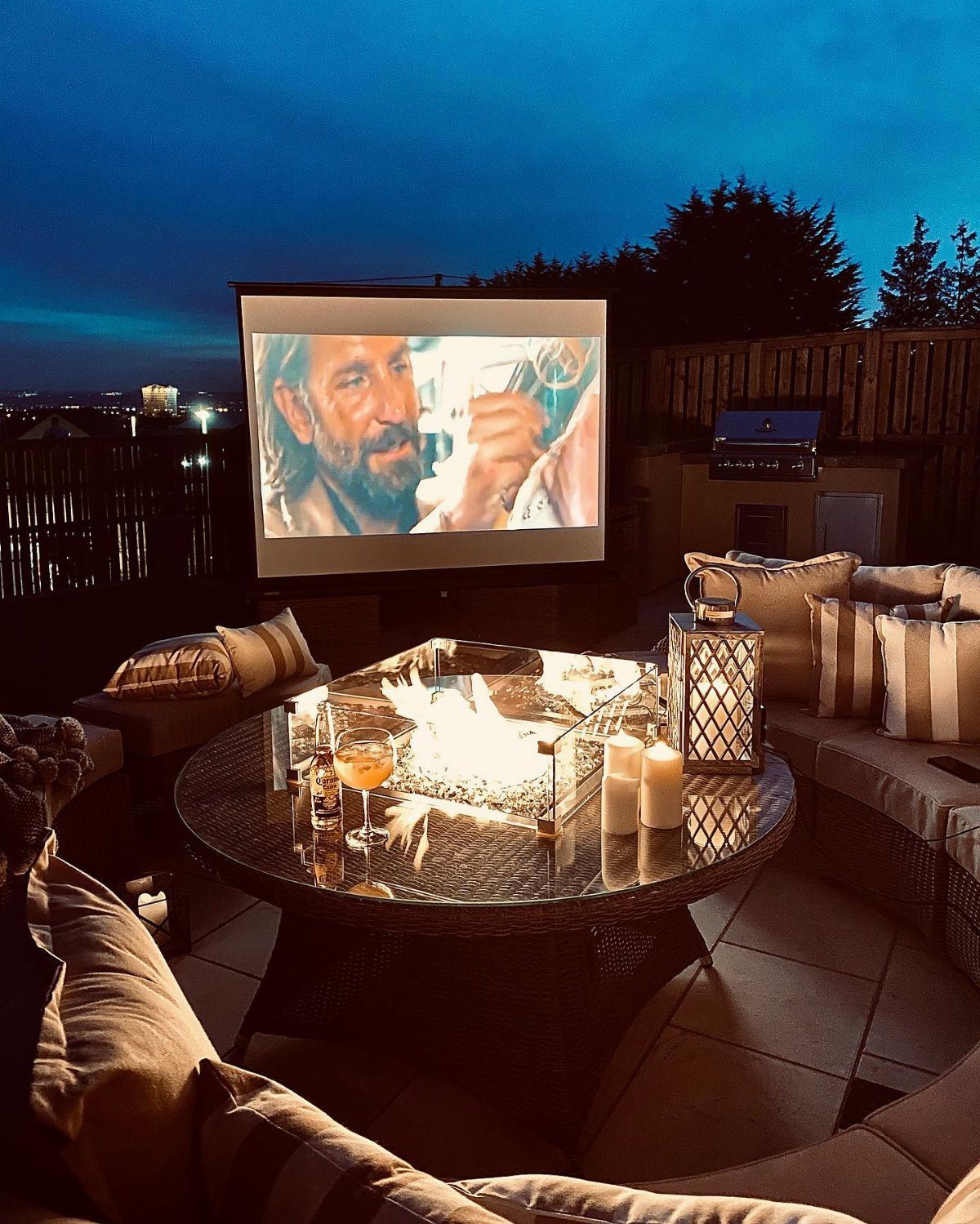 Outdoor patio setup at night with a projection screen, fire pit table, and lounge seating.
