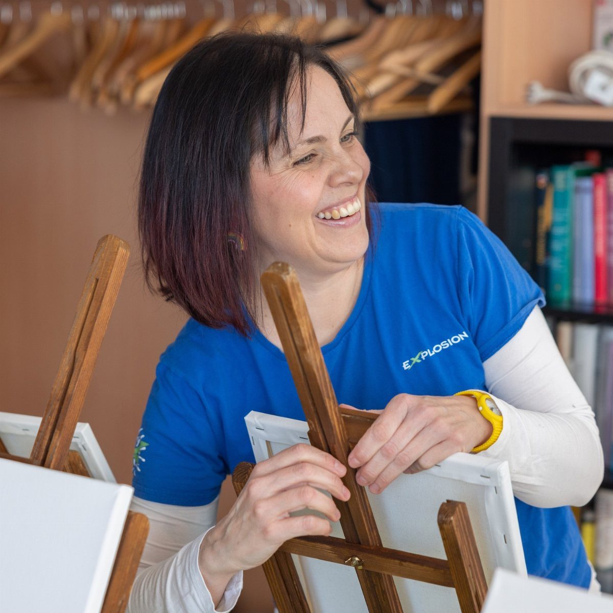 A smiling woman in a blue shirt working with small wooden easels and white canvases.